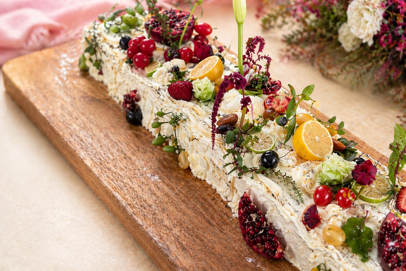 A long cream cake on a wooden board, layered with fresh berries, citrus and edible flowers